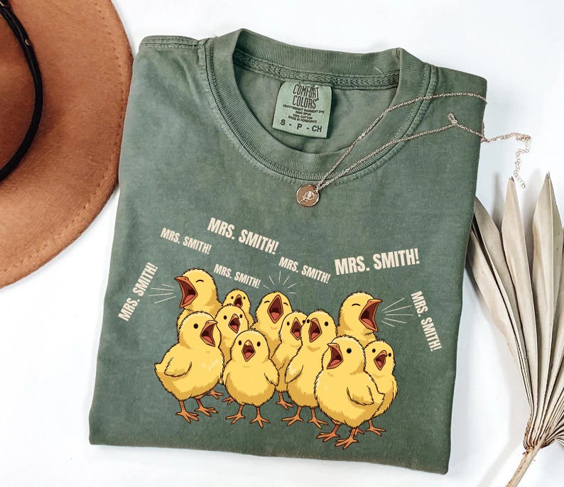 May include: A sage green t-shirt with a graphic of yellow chicks yelling "MRS. SMITH!". The shirt has a crew neck and short sleeves. A silver necklace with a pendant is draped on the shirt.