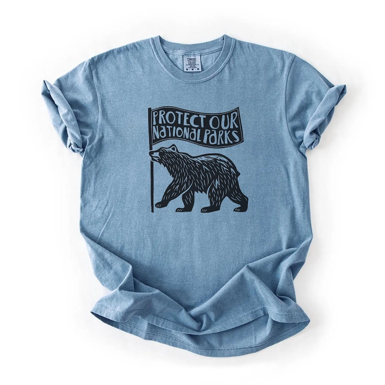 May include: A blue t-shirt with a black bear holding a flag that reads 'Protect Our National Parks'.