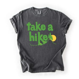 May include: Dark gray t-shirt with the words "take a hike" in bright green. The design includes a green mountain, a yellow sun, and hiking boots. The shirt has rolled-up sleeves and a crew neck.