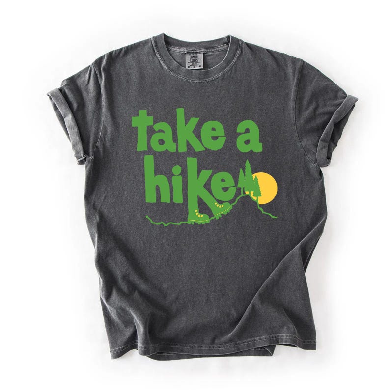 May include: Dark gray t-shirt with the words "take a hike" in bright green. The design includes a green mountain, a yellow sun, and hiking boots. The shirt has rolled-up sleeves and a crew neck.