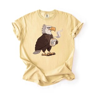 Puede incluir: Camiseta amarillo claro con un &aacute;guila de dibujos animados sosteniendo una taza de caf&eacute;. El &aacute;guila es marr&oacute;n, blanca y gris, con una expresi&oacute;n severa. La taza dice "IS HE DEAD YET?" en texto negro. La camiseta tiene mangas enrolladas.