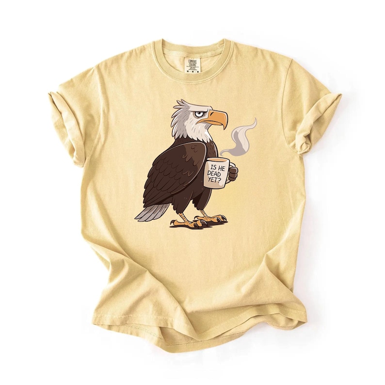 Puede incluir: Camiseta amarillo claro con un &aacute;guila de dibujos animados sosteniendo una taza de caf&eacute;. El &aacute;guila es marr&oacute;n, blanca y gris, con una expresi&oacute;n severa. La taza dice "IS HE DEAD YET?" en texto negro. La camiseta tiene mangas enrolladas.