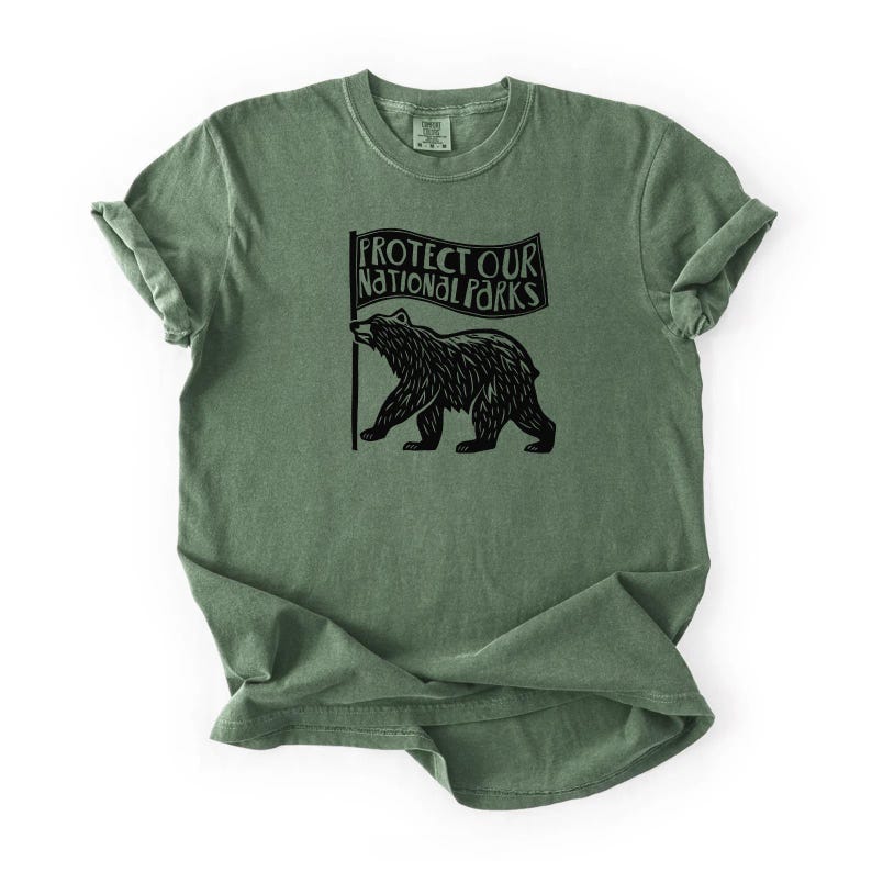 May include: Green t-shirt with rolled sleeves. It features a black graphic of a bear holding a flag with the text "PROTECT OUR NATIONAL PARKS". The shirt has a vintage, worn appearance.