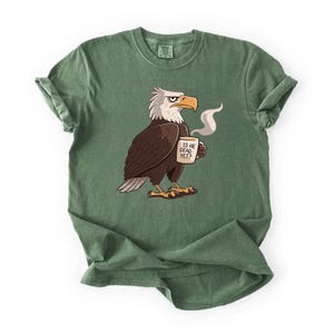 Puede incluir: Camiseta verde con una ilustraci&oacute;n de un &aacute;guila de dibujos animados. El &aacute;guila tiene la cabeza blanca, el cuerpo marr&oacute;n y el pico y las patas amarillas. Sostiene una taza con el texto "IS HE DEAD YET?" y vapor saliendo. La camiseta tiene las mangas enrolladas.
