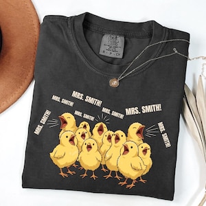 May include: Dark gray Comfort Colors t-shirt featuring a graphic of yellow chicks with open beaks, shouting "MRS. SMITH!". The shirt has a crew neck and short sleeves. A silver necklace with a pendant is also visible.