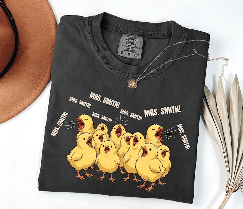 May include: Dark gray Comfort Colors t-shirt featuring a graphic of yellow chicks with open beaks, shouting "MRS. SMITH!". The shirt has a crew neck and short sleeves. A silver necklace with a pendant is also visible.