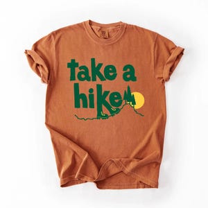 May include: A rust-colored t-shirt with the words "take a hike" in green block letters. The design includes green hiking boots, trees, and a yellow sun over a mountain. The shirt has rolled-up sleeves.