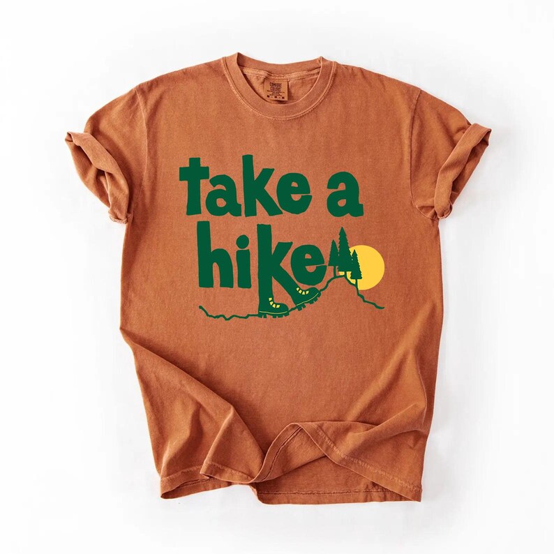 May include: A rust-colored t-shirt with the words "take a hike" in green block letters. The design includes green hiking boots, trees, and a yellow sun over a mountain. The shirt has rolled-up sleeves.