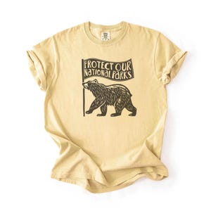 May include: A tan t-shirt featuring a black bear graphic holding a flag with the text "Protect Our National Parks." The shirt is made of a soft material and has rolled-up sleeves.