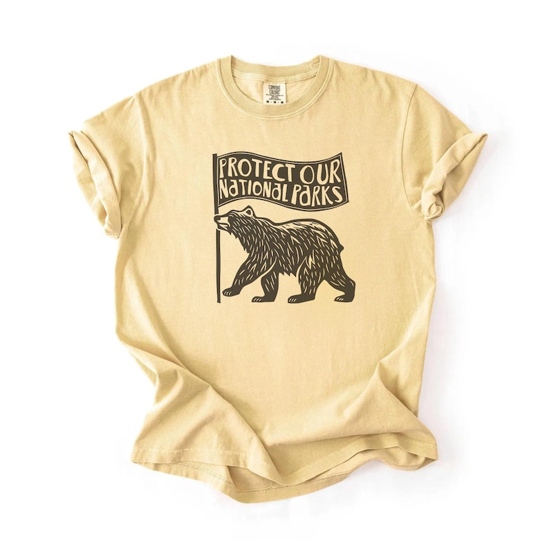 May include: A tan t-shirt featuring a black bear graphic holding a flag with the text "Protect Our National Parks." The shirt is made of a soft material and has rolled-up sleeves.