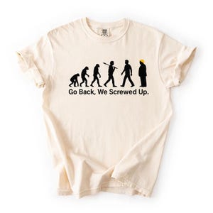 Puede incluir: Camiseta color crema con un gráfico de evolución en silueta negra. El gráfico representa la evolución del hombre, culminando en una figura con peinado amarillo. El texto "Go Back, We Screwed Up" está debajo.