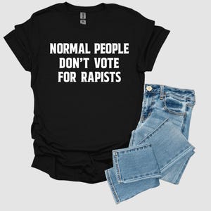 May include: Black t-shirt with the white text "NORMAL PEOPLE DON'T VOTE FOR RAPISTS." The shirt is paired with light blue denim jeans. The t-shirt is folded, and the jeans are neatly stacked.