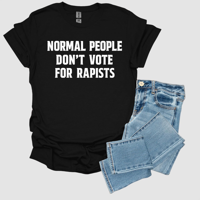 May include: Black t-shirt with the white text "NORMAL PEOPLE DON'T VOTE FOR RAPISTS." The shirt is paired with light blue denim jeans. The t-shirt is folded, and the jeans are neatly stacked.