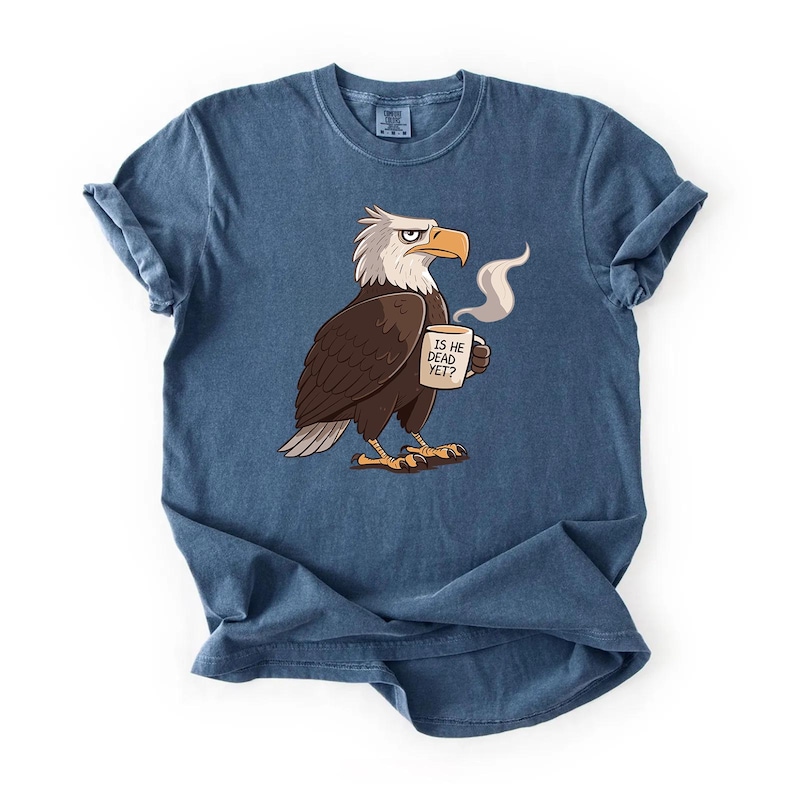 Puede incluir: Camiseta azul con un dise&ntilde;o de &aacute;guila de dibujos animados sosteniendo una taza humeante. El &aacute;guila tiene la cabeza blanca y el cuerpo marr&oacute;n. La taza dice "IS HE DEAD YET?". La camiseta tiene mangas enrolladas.