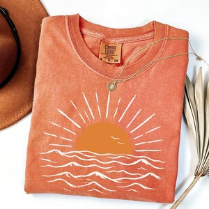 May include: A terracotta-colored short-sleeve t-shirt featuring a graphic of a solid orange sun rising over white stylized ocean waves. The shirt has a crew neckline and a Comfort Colors brand label printed on the inside of the collar area.