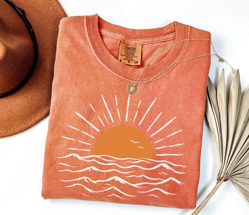 May include: A terracotta-colored short-sleeve t-shirt featuring a graphic of a solid orange sun rising over white stylized ocean waves. The shirt has a crew neckline and a Comfort Colors brand label printed on the inside of the collar area.
