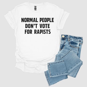 May include: White t-shirt with the text "NORMAL PEOPLE DON'T VOTE FOR RAPISTS" in black. The shirt is paired with blue denim jeans. The t-shirt has a crew neck and short sleeves.