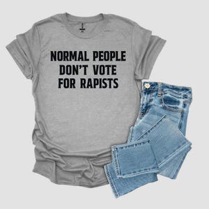 May include: A gray t-shirt with the text "NORMAL PEOPLE DON'T VOTE FOR RAPISTS" in black, paired with folded blue denim jeans. The shirt has a crew neck and short sleeves.