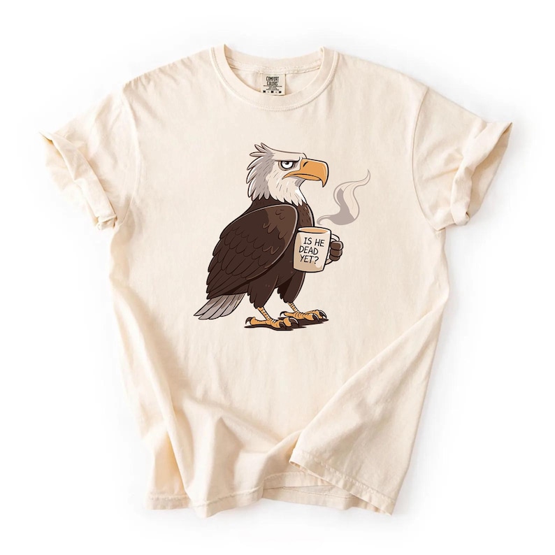 Puede incluir: Camiseta color crema con un &aacute;guila de dibujos animados sosteniendo una taza con la frase "IS HE DEAD YET?". El &aacute;guila es marr&oacute;n, blanca y gris, con pico y garras amarillas. La camiseta tiene las mangas enrolladas. El dise&ntilde;o est&aacute; impreso.