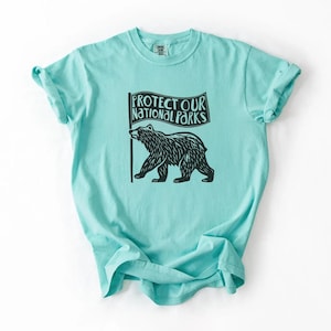 May include: A light turquoise t-shirt with a black graphic of a bear holding a flag. The flag reads "PROTECT OUR NATIONAL PARKS." The shirt has short sleeves and a crew neck. The fabric appears soft and comfortable.
