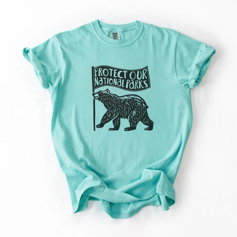 May include: A light turquoise t-shirt with a black graphic of a bear holding a flag. The flag reads "PROTECT OUR NATIONAL PARKS." The shirt has short sleeves and a crew neck. The fabric appears soft and comfortable.