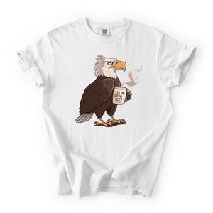 Puede incluir: Camiseta blanca con un dise&ntilde;o de &aacute;guila calva de dibujos animados sosteniendo una taza de caf&eacute;. El &aacute;guila tiene plumas marrones, cabeza blanca y pico y patas amarillas. La taza dice "IS HE DEAD YET?"