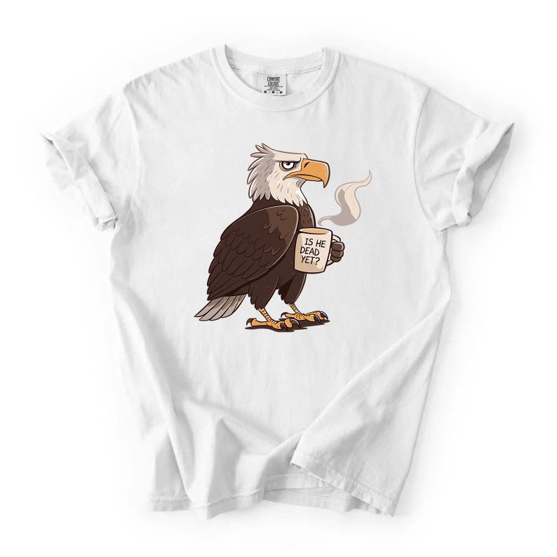 Puede incluir: Camiseta blanca con un dise&ntilde;o de &aacute;guila calva de dibujos animados sosteniendo una taza de caf&eacute;. El &aacute;guila tiene plumas marrones, cabeza blanca y pico y patas amarillas. La taza dice "IS HE DEAD YET?"