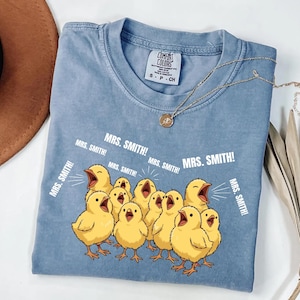 May include: A light blue Comfort Colors t-shirt featuring a graphic of yellow chicks with open beaks, and the text "MRS. SMITH!". A gold necklace with a pendant is draped on the shirt.
