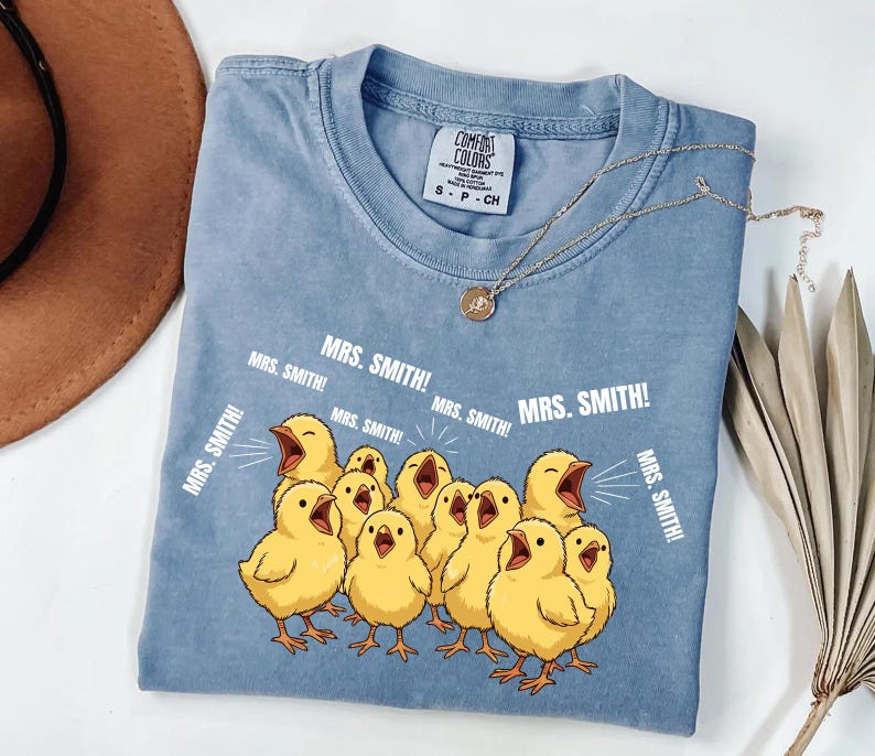 May include: A light blue Comfort Colors t-shirt featuring a graphic of yellow chicks with open beaks, and the text "MRS. SMITH!". A gold necklace with a pendant is draped on the shirt.