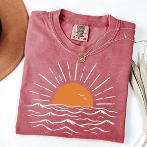 May include: A dusty rose short-sleeve t-shirt featuring a graphic of an orange sun rising over white stylized ocean waves. The shirt has a crew neckline and a Comfort Colors brand label on the inside collar. A thin gold chain necklace rests on top.