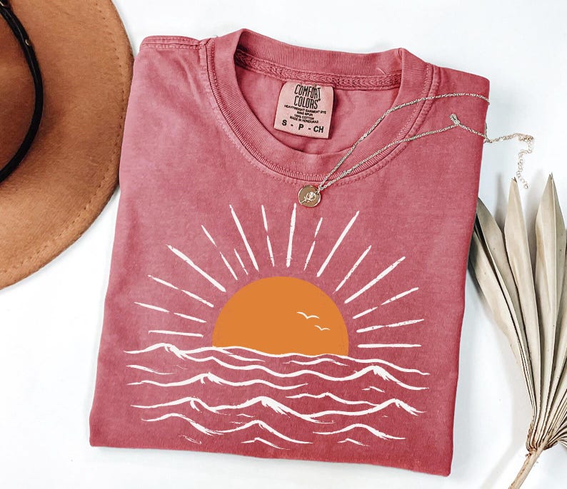 May include: A dusty rose short-sleeve t-shirt featuring a graphic of an orange sun rising over white stylized ocean waves. The shirt has a crew neckline and a Comfort Colors brand label on the inside collar. A thin gold chain necklace rests on top.