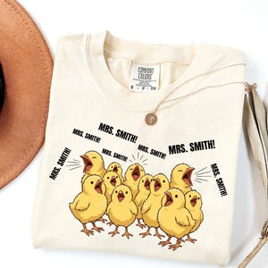 May include: Cream-colored Comfort Colors t-shirt featuring a graphic of cartoon yellow chicks with open beaks, shouting "MRS. SMITH!". The shirt has a small tag and is paired with a gold necklace. A brown hat and dried plant are in the background.
