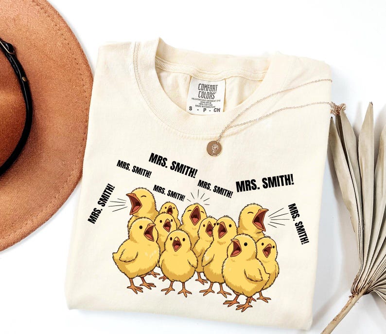 May include: Cream-colored Comfort Colors t-shirt featuring a graphic of cartoon yellow chicks with open beaks, shouting "MRS. SMITH!". The shirt has a small tag and is paired with a gold necklace. A brown hat and dried plant are in the background.