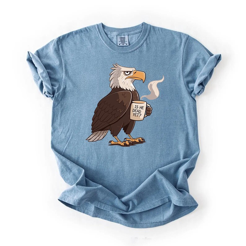 Puede incluir: Camiseta azul claro con un &aacute;guila de dibujos animados sosteniendo una taza humeante. El &aacute;guila tiene una expresi&oacute;n seria y la taza dice "IS HE DEAD YET?". La camiseta es de color liso, con cuello redondo y mangas cortas.