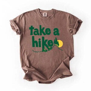 May include: Brown t-shirt with the words "take a hike" in green, along with a graphic of hiking boots, a mountain, and a yellow sun. The shirt has a crew neck and short sleeves.