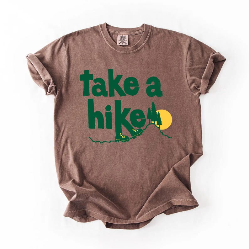 May include: Brown t-shirt with the words "take a hike" in green, along with a graphic of hiking boots, a mountain, and a yellow sun. The shirt has a crew neck and short sleeves.