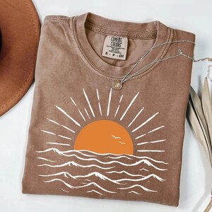 May include: A brown short-sleeve t-shirt featuring a graphic of an orange sun rising over white stylized ocean waves. The shirt has a crew neckline and a Comfort Colors brand label on the inside collar. A silver chain necklace rests on the fabric.