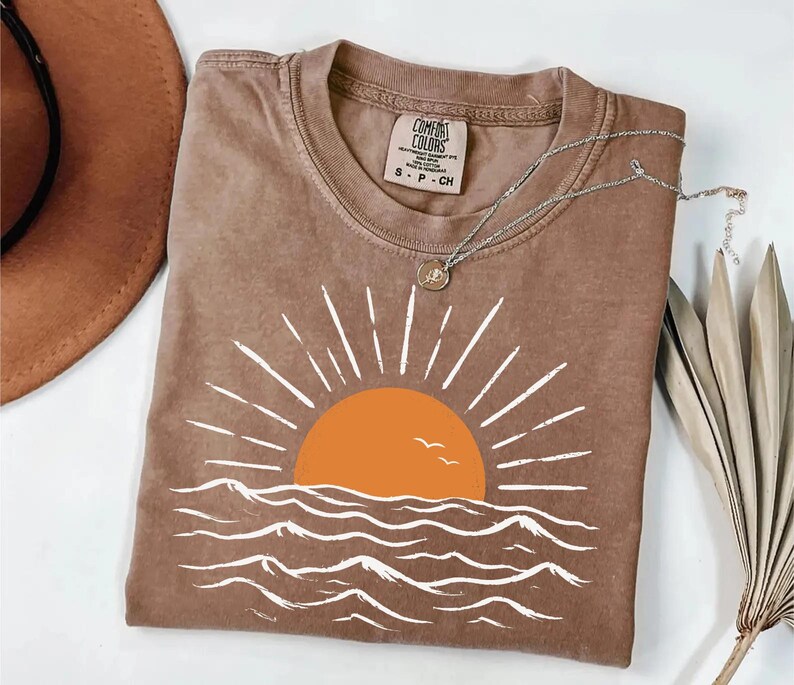 May include: A brown short-sleeve t-shirt featuring a graphic of an orange sun rising over white stylized ocean waves. The shirt has a crew neckline and a Comfort Colors brand label on the inside collar. A silver chain necklace rests on the fabric.