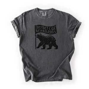 May include: Dark grey t-shirt with rolled sleeves. The shirt displays a black graphic of a bear holding a flag with the text "PROTECT OUR NATIONAL PARKS". The t-shirt has a vintage, worn appearance.
