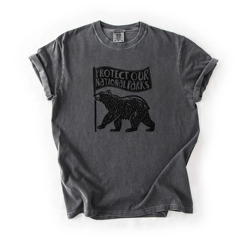 May include: Dark grey t-shirt with rolled sleeves. The shirt displays a black graphic of a bear holding a flag with the text "PROTECT OUR NATIONAL PARKS". The t-shirt has a vintage, worn appearance.