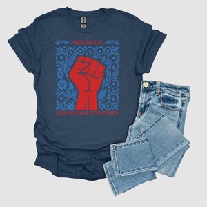 May include: A navy blue t-shirt with a red fist graphic inside a blue floral design. The text "ABOLISH ICE" and "KEEP FAMILIES TOGETHER" are printed on the shirt. Light blue denim jeans are folded next to the shirt.