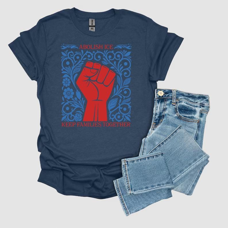 May include: A navy blue t-shirt with a red fist graphic inside a blue floral design. The text "ABOLISH ICE" and "KEEP FAMILIES TOGETHER" are printed on the shirt. Light blue denim jeans are folded next to the shirt.