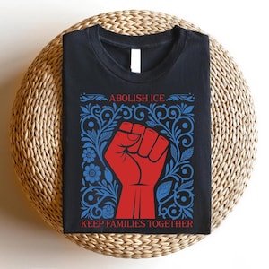 May include: Black t-shirt with a red fist graphic inside a blue floral design. The text "ABOLISH ICE" is above the fist, and "KEEP FAMILIES TOGETHER" is below. The shirt is displayed on a woven, circular surface.