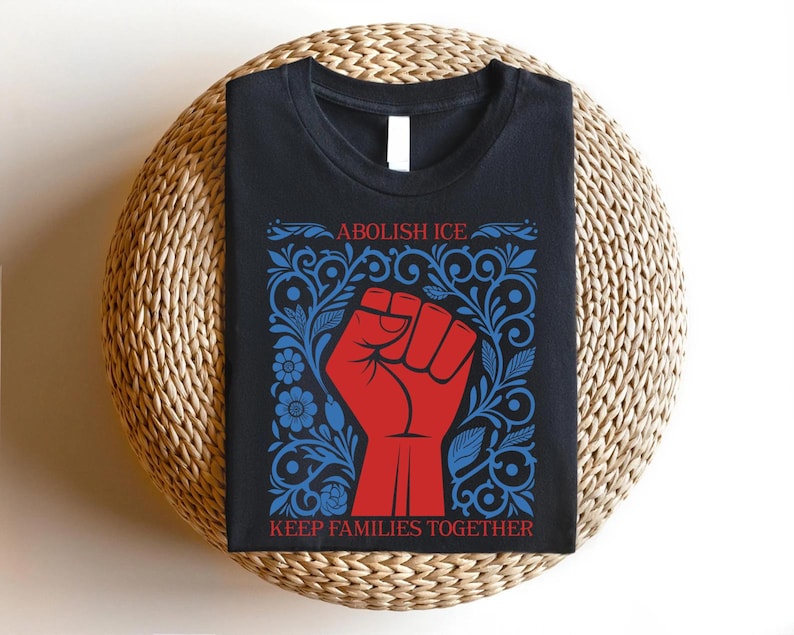 May include: Black t-shirt with a red fist graphic inside a blue floral design. The text "ABOLISH ICE" is above the fist, and "KEEP FAMILIES TOGETHER" is below. The shirt is displayed on a woven, circular surface.