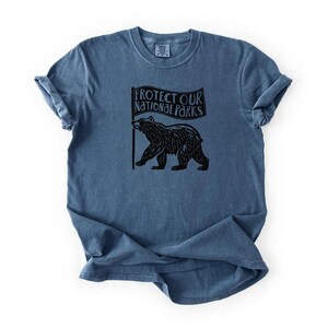 May include: A blue t-shirt featuring a black bear graphic holding a flag with the text "PROTECT OUR NATIONAL PARKS." The shirt has a crew neck and short sleeves.