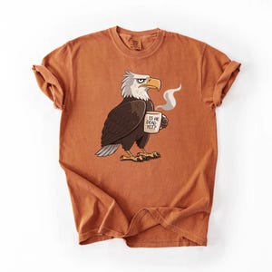 Puede incluir: Camiseta naranja quemado con un &aacute;guila de dibujos animados sosteniendo una taza de caf&eacute;. El &aacute;guila tiene plumas blancas y marrones y un pico amarillo. La taza tiene el texto "IS HE DEAD YET?". La camiseta tiene las mangas enrolladas.