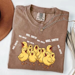 May include: Brown Comfort Colors t-shirt featuring a graphic of yellow chicks with open beaks, shouting "MRS. SMITH!". The shirt has a crew neck and a small tag. A silver necklace with a pendant is draped across the shirt.