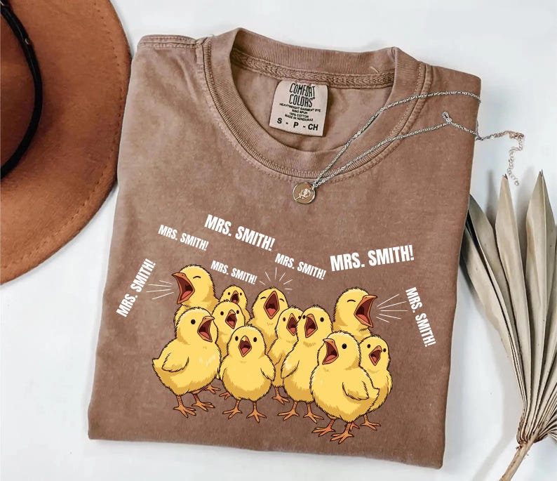 May include: Brown Comfort Colors t-shirt featuring a graphic of yellow chicks with open beaks, shouting "MRS. SMITH!". The shirt has a crew neck and a small tag. A silver necklace with a pendant is draped across the shirt.