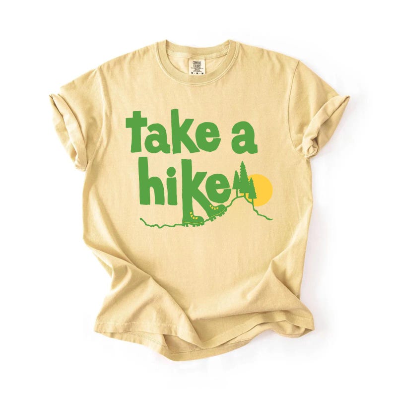 May include: A light yellow t-shirt with the words "take a hike" in green block letters. The design includes green hiking boots, a mountain, and a yellow sun. The shirt is made of a soft material and has rolled-up sleeves.