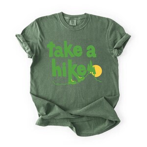 May include: Green t-shirt with the phrase "take a hike" in bright green letters. The design includes hiking boots, a mountain, and a yellow sun. The shirt is a solid green color.